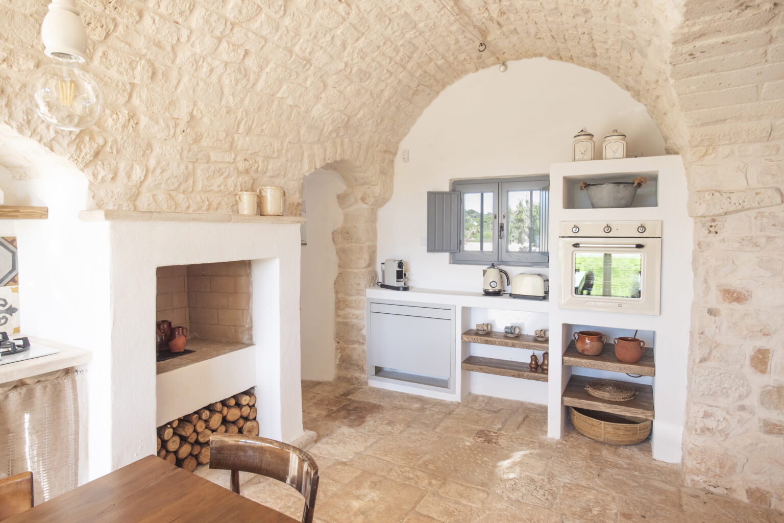 Cozy rustic kitchen featuring arched stone walls, a wooden dining chair, a fireplace with stacked firewood, and a modern kitchen setup with a white cabinet and gray window. Natural light illuminates the space, enhancing its warm, inviting atmosphere. | Only on SopranoVillas