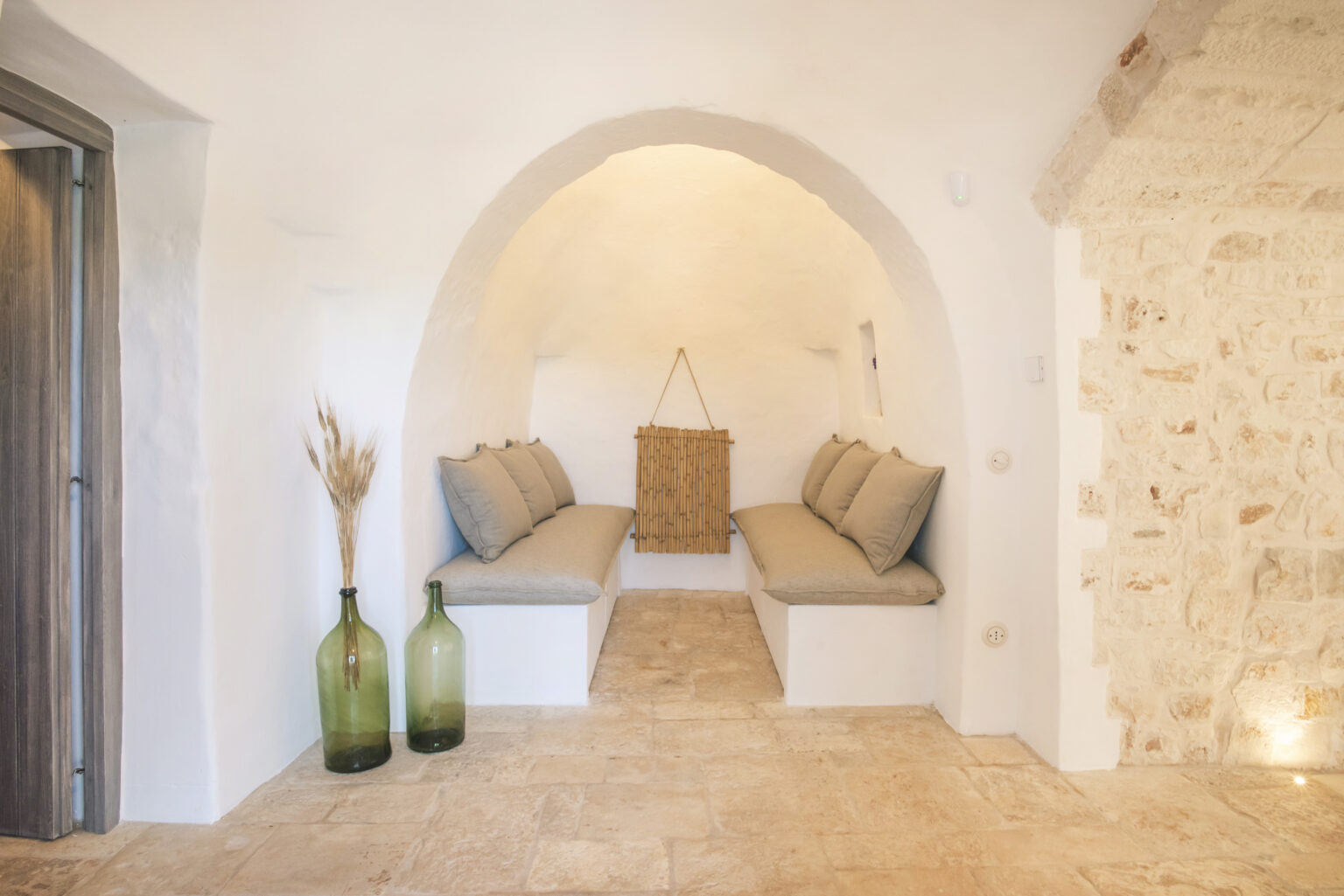 Interior view of a cozy, minimalist seating area featuring two built-in benches with soft cushions, an arched entrance, and decorative glass bottles. The walls are a mix of smooth white and rustic stone, creating a warm and inviting atmosphere. | Only on SopranoVillas