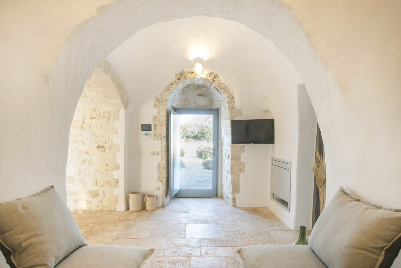 Interior view of a cozy, modern living space featuring arched stone walls, a light-colored sofa, and a large glass door leading to an outdoor area. Natural light illuminates the room, enhancing the rustic charm of the design. | Only on SopranoVillas