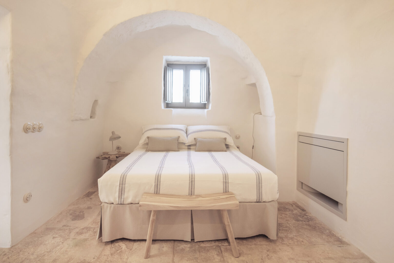 Cozy bedroom with an arched ceiling, featuring a double bed with striped bedding, a wooden bench, and a window letting in natural light, set in a minimalist white interior. | Only on SopranoVillas