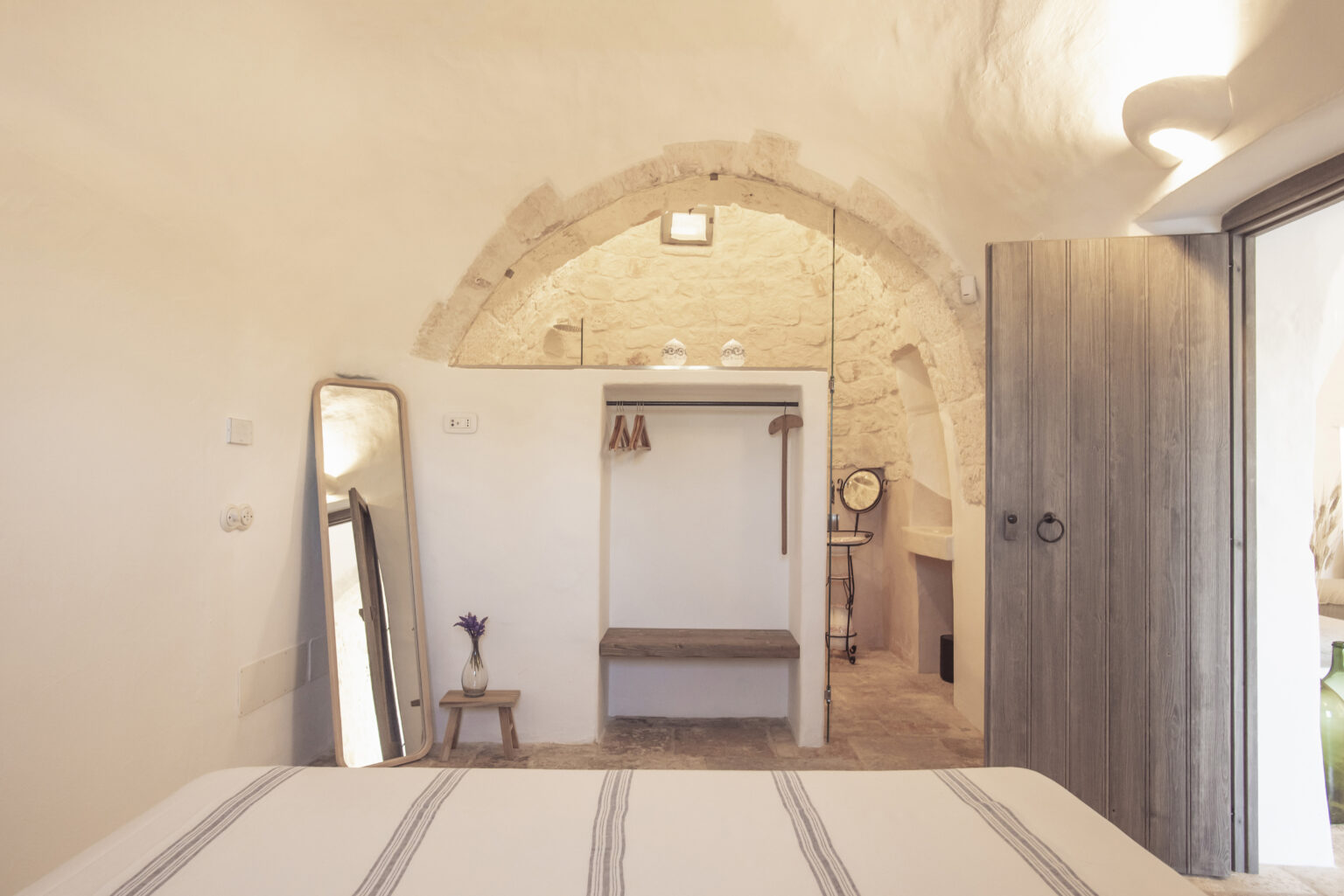 Cozy bedroom interior featuring a stone arch ceiling, minimalist decor, a large mirror, and a wooden bench for seating. The room is well-lit, highlighting the natural textures and warm tones of the materials used. | Only on SopranoVillas
