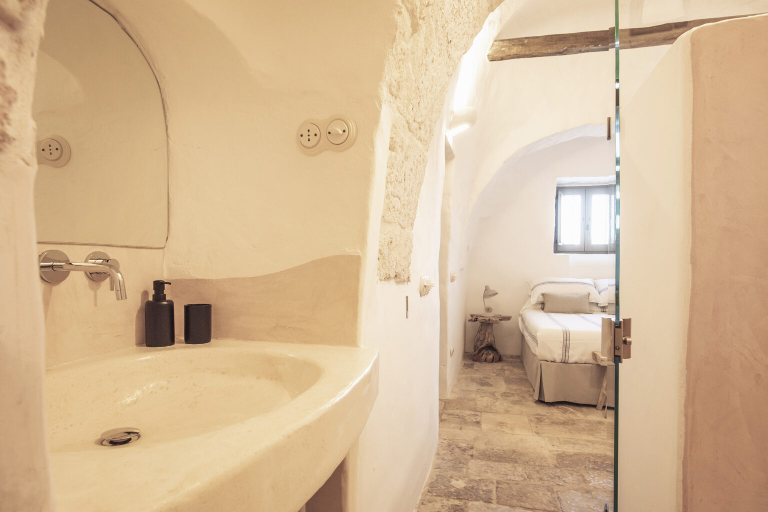 Modern bathroom with a minimalist design featuring a curved sink, natural stone flooring, and an adjoining bedroom visible through a glass shower door. The space is illuminated by natural light from a window, creating a warm and inviting atmosphere. | Only on SopranoVillas