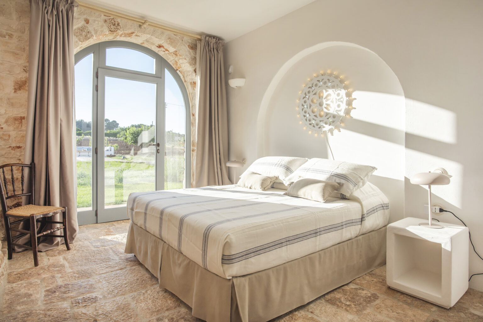 Cozy bedroom featuring a large window with natural light, a comfortable bed with neutral bedding, and minimalist decor, set in a stylish, rustic interior. | Only on SopranoVillas