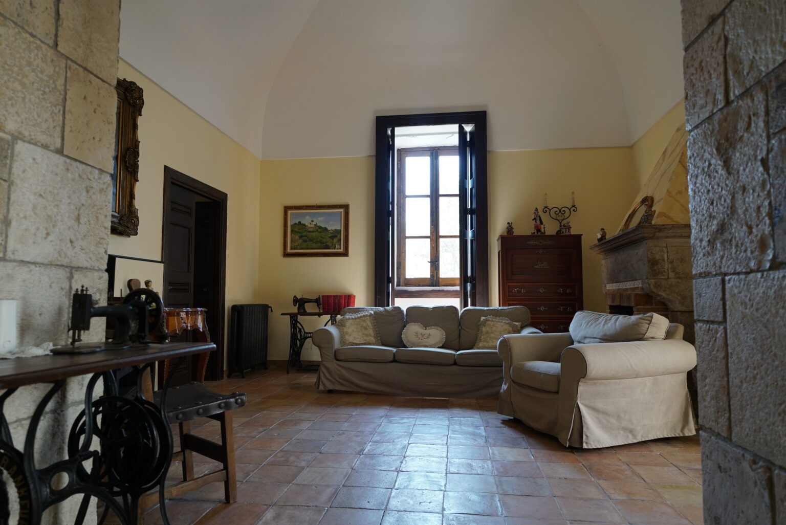 Cozy living room featuring a beige sofa, vintage furniture, and a large window allowing natural light, with stone walls and terracotta flooring. | Only on SopranoVillas