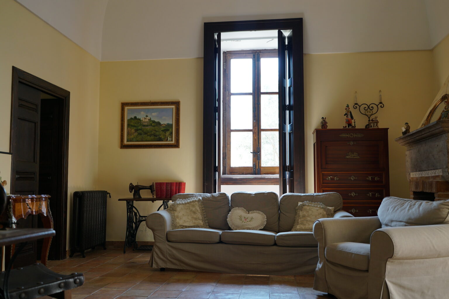 Cozy living room interior featuring a beige sofa, large window with wooden shutters, a decorative painting, and a wooden cabinet, creating a warm and inviting atmosphere. | Only on SopranoVillas
