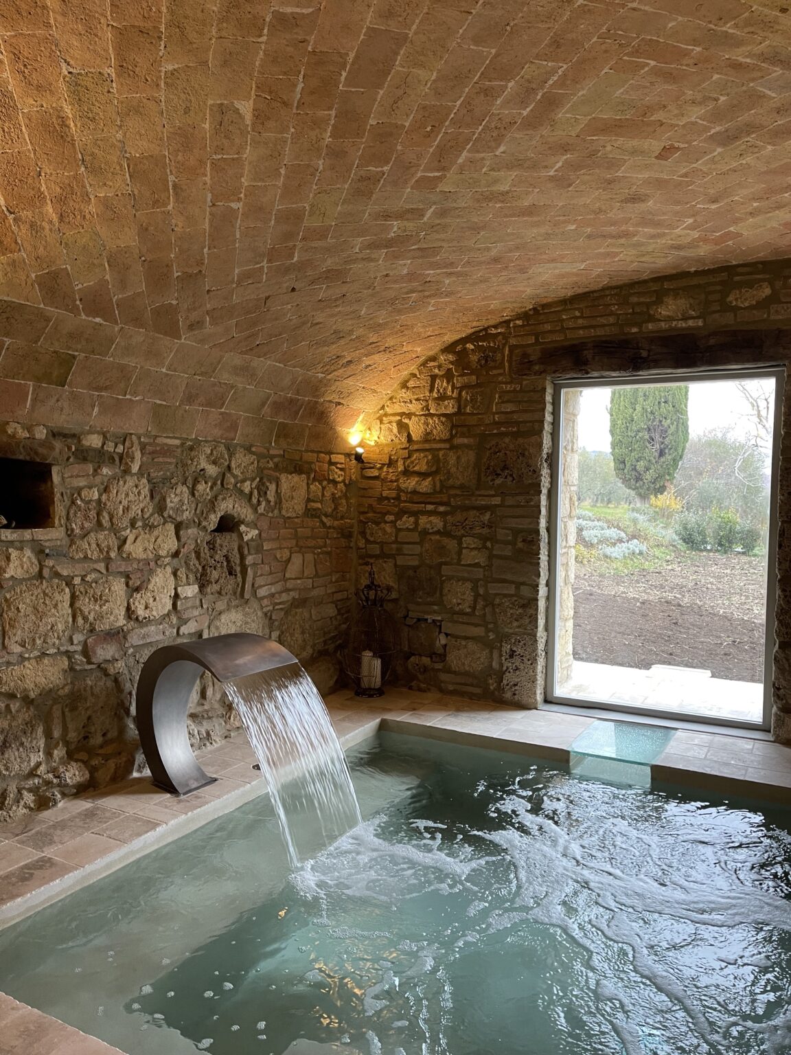 Interior view of a serene spa with a stone wall, featuring a modern water fountain and a large window overlooking a landscaped garden, creating a tranquil atmosphere for relaxation. | Only on SopranoVillas