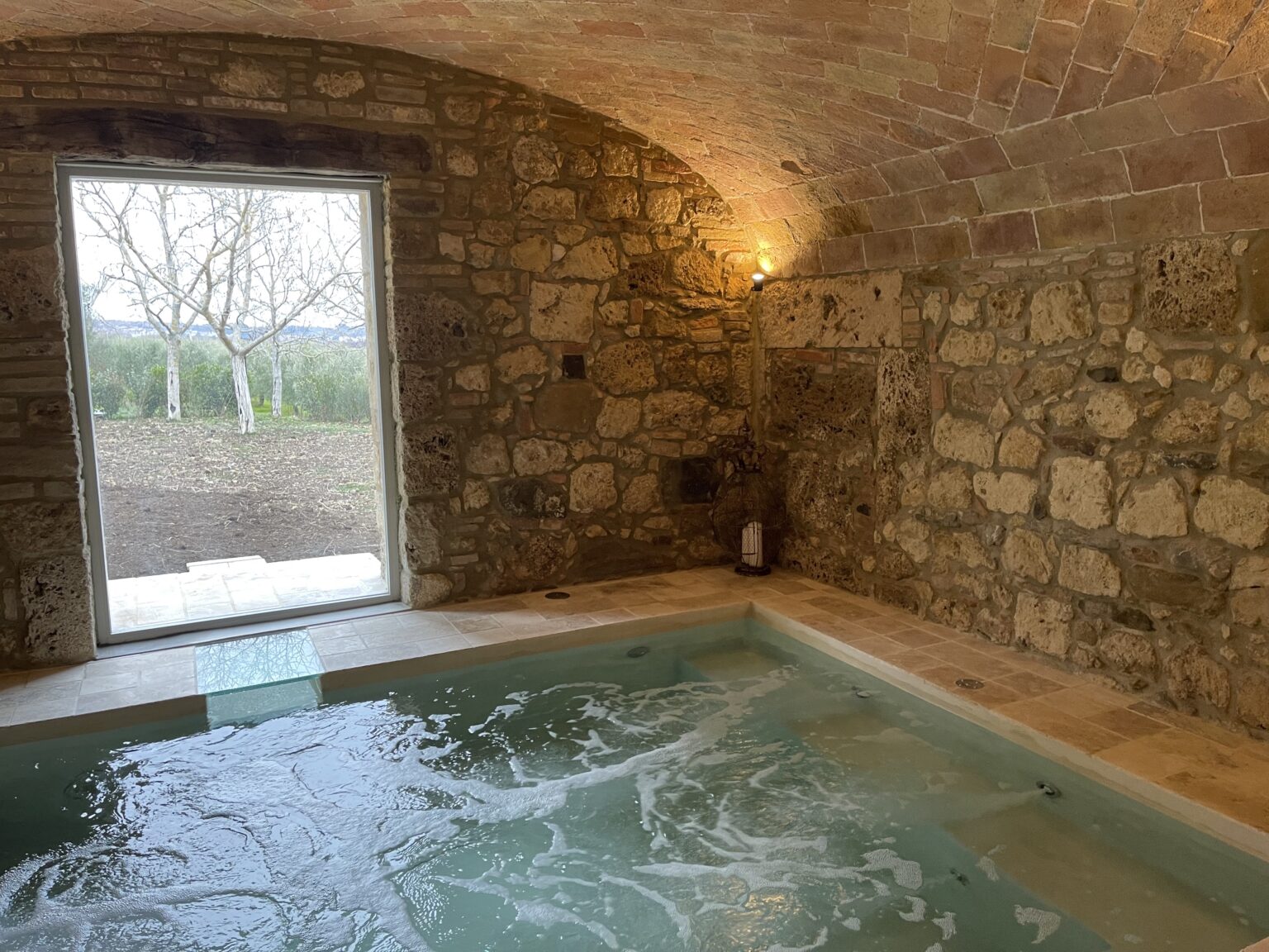 Indoor spa area featuring a stone wall, a jacuzzi, and a large window offering views of the outdoors. The warm lighting creates a relaxing atmosphere perfect for wellness and relaxation. | Only on SopranoVillas