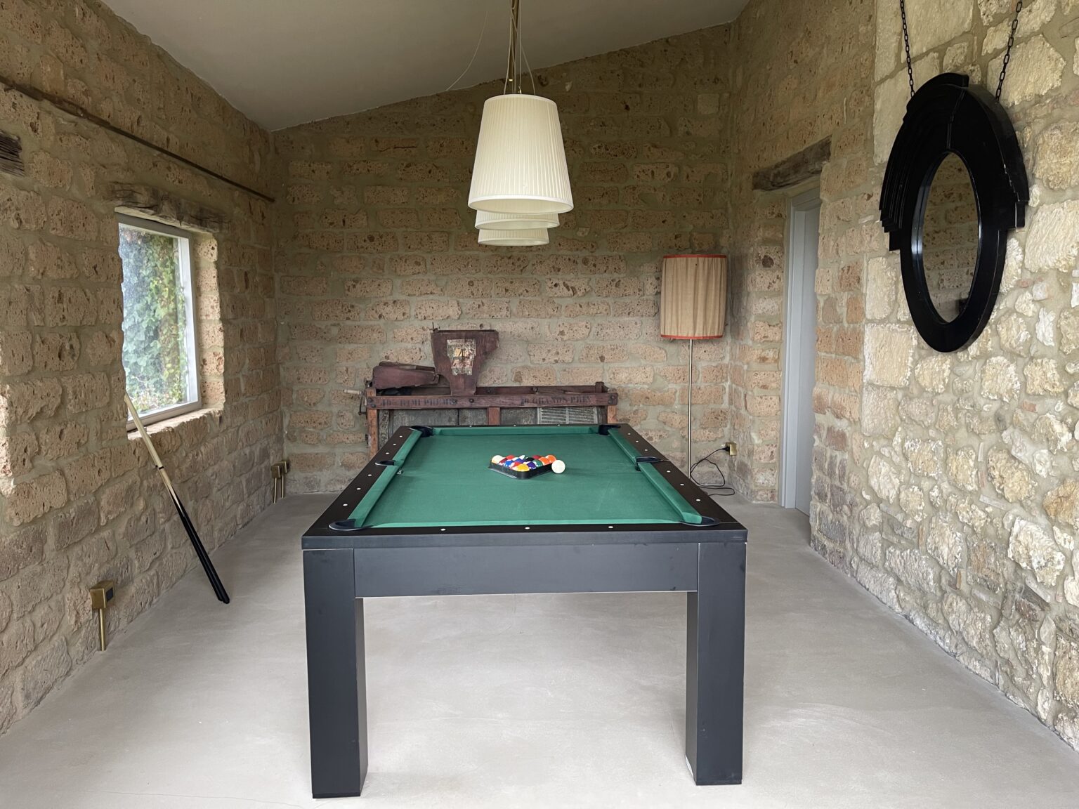 Interior of a rustic game room featuring a green pool table, stone walls, and a pendant light. A wooden chair and a decorative black circle on the wall add to the cozy atmosphere. | Only on SopranoVillas