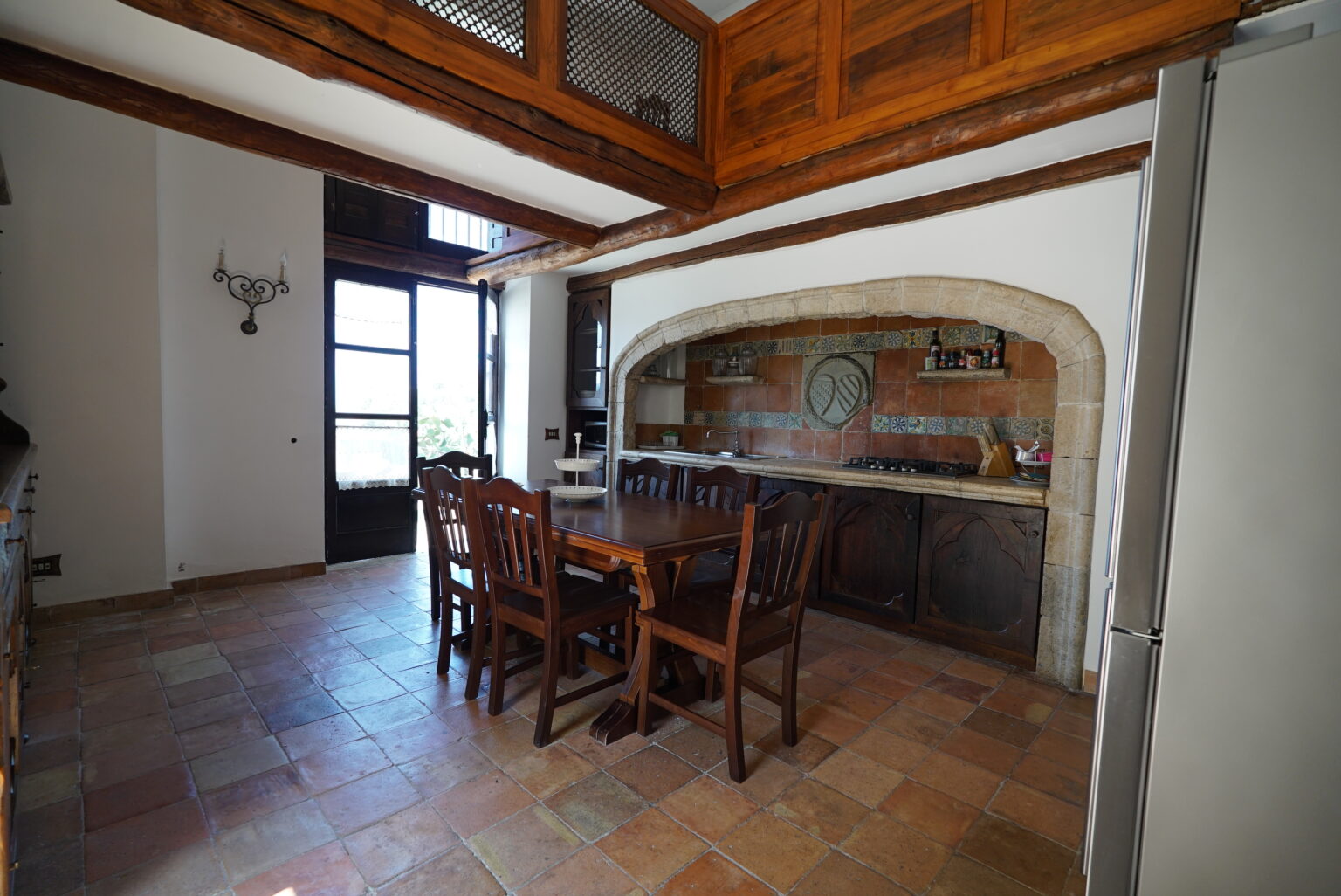 Spacious kitchen with wooden beams, featuring a dining table and chairs, stone archway, and modern appliances, illuminated by natural light from large windows. | Only on SopranoVillas
