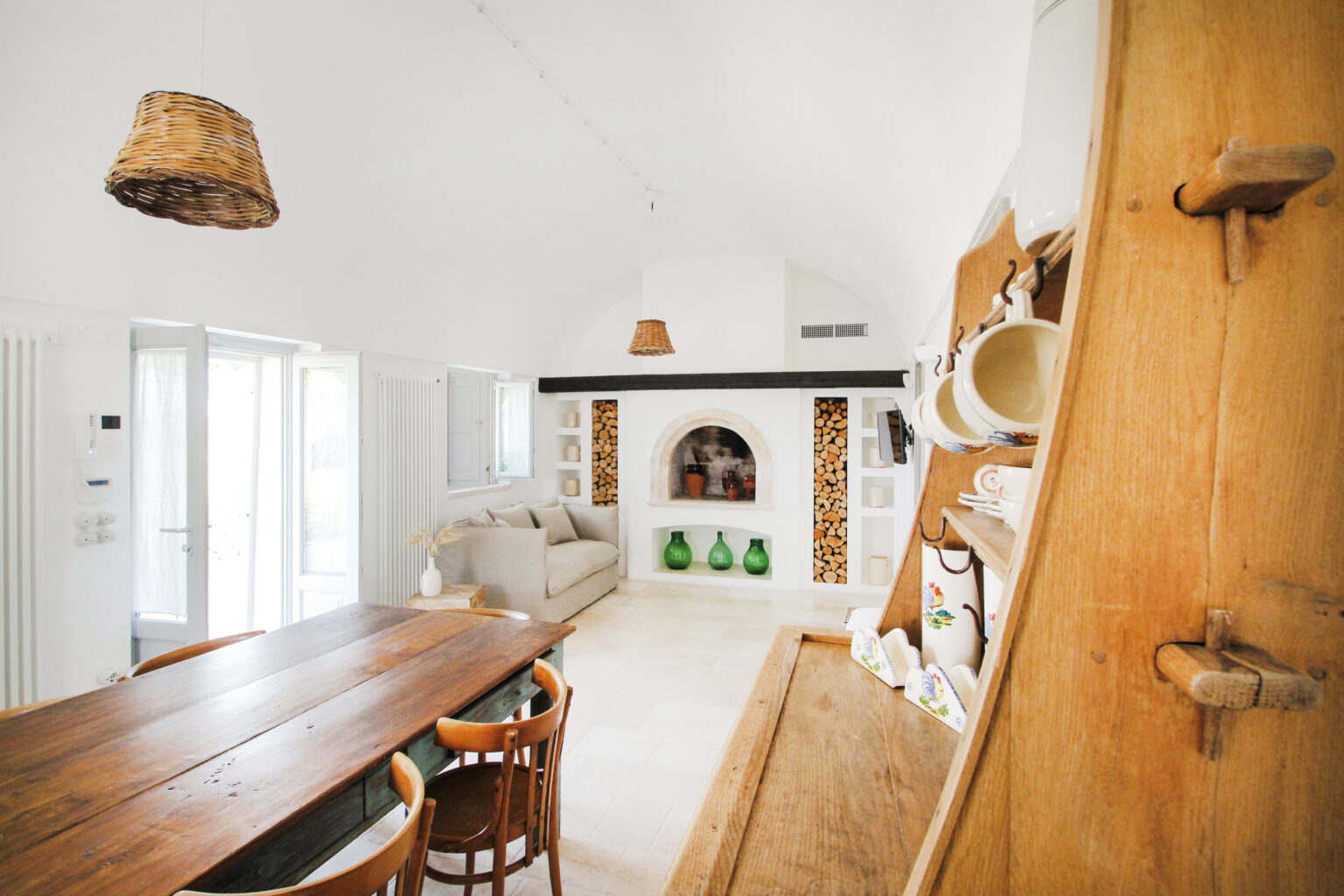 Bright and airy interior of a modern living room featuring a wooden dining table, comfortable sofa, decorative fireplace, and natural light streaming through large windows. The decor includes earthy tones and stylish accents, creating a welcoming atmosphere. | Only on SopranoVillas