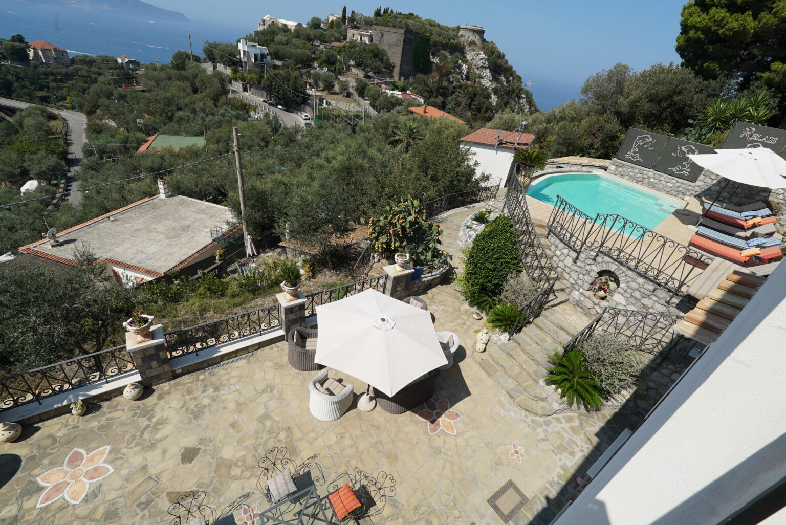 Scenic view of a villa terrace featuring a swimming pool, sun loungers, and shaded seating area surrounded by lush greenery and hills, with a glimpse of the ocean in the background. | Only on SopranoVillas