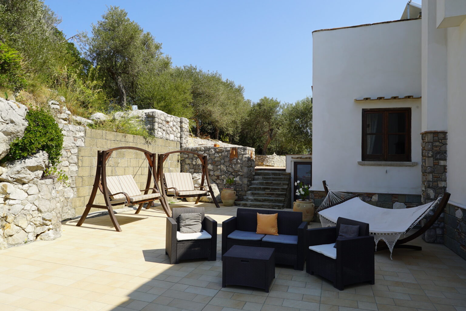 Outdoor patio area featuring wooden swings, a hammock, and modern seating arrangements surrounded by lush greenery and stone walls, ideal for relaxation and entertaining. | Only on SopranoVillas