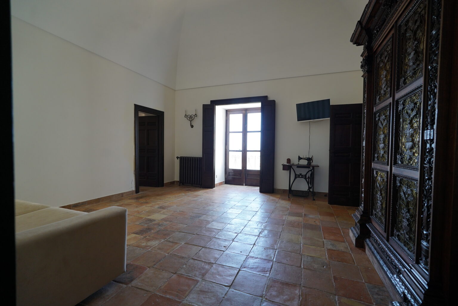 Spacious interior of a stylish room featuring terracotta tiled flooring, large windows with wooden frames, a vintage sewing machine on a small table, and an ornate wooden cabinet, creating a blend of modern and traditional decor. | Only on SopranoVillas