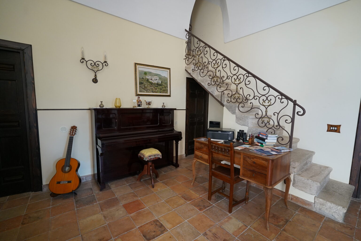 Cozy home interior featuring a wooden piano, a vintage desk with office supplies, a guitar, and an elegant staircase with wrought iron railing. Warm tile flooring enhances the inviting atmosphere. | Only on SopranoVillas