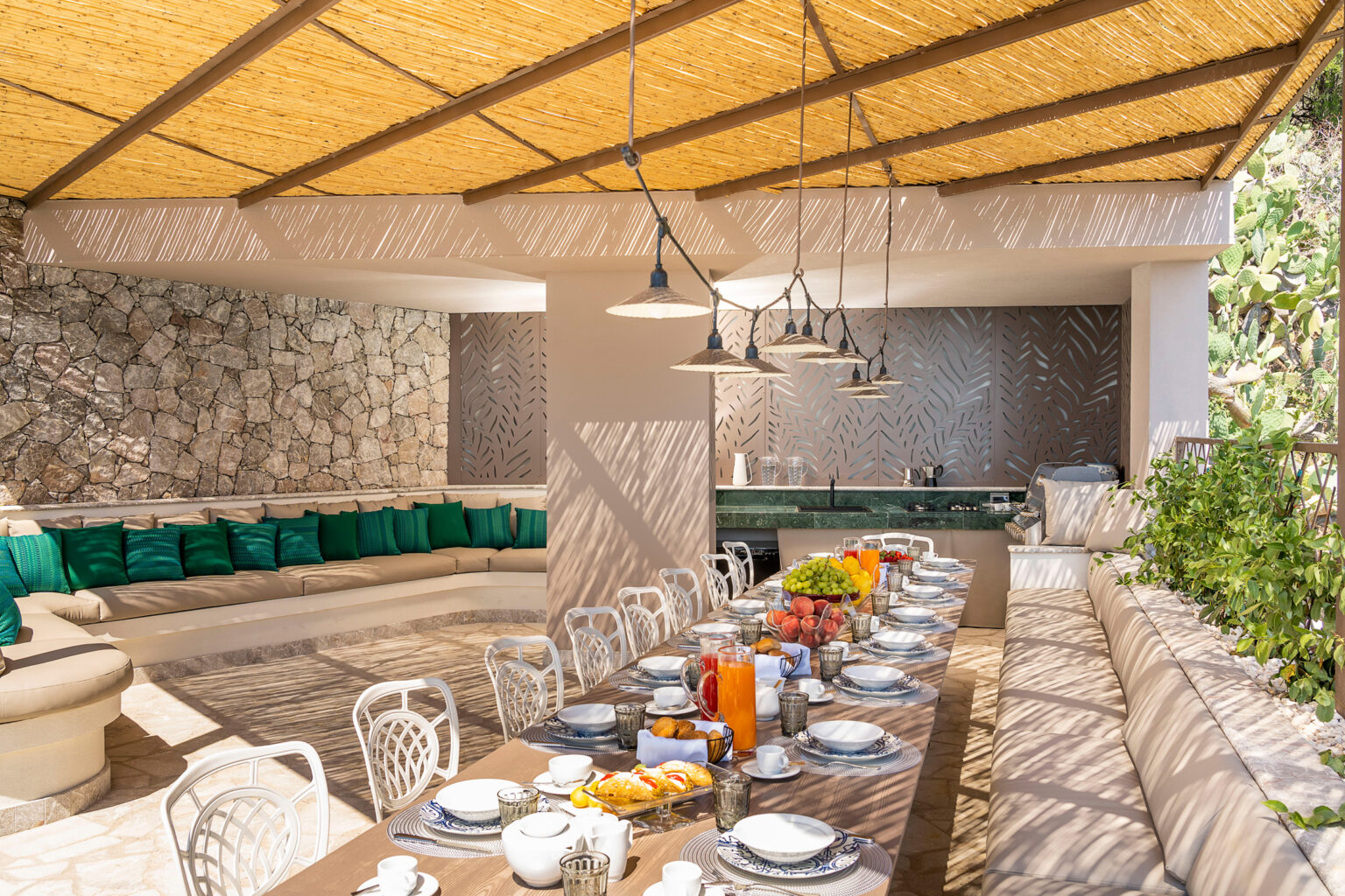 Outdoor dining area with a long wooden table set for a meal, surrounded by green cushions and a modern kitchen. Natural light filters through a bamboo roof, highlighting the stone walls and lush greenery. | Only on SopranoVillas