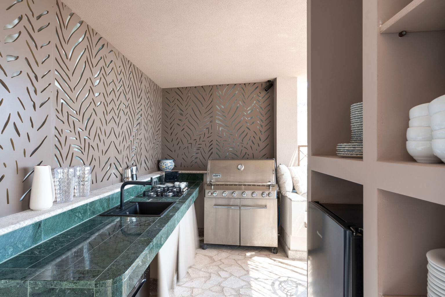 Modern outdoor kitchen featuring a sleek granite countertop, built-in grill, and stylish patterned wall. The space is designed for entertaining, complete with storage shelves and a compact refrigerator, ideal for outdoor cooking and gatherings. | Only on SopranoVillas