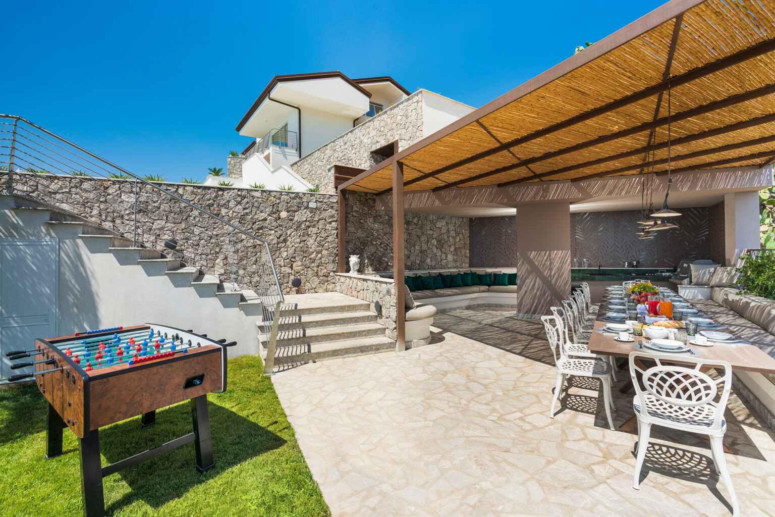 Outdoor entertainment area featuring a foosball table, a dining setup under a shaded patio, and a luxurious pool in a modern villa. Bright sunny skies enhance the inviting atmosphere. | Only on SopranoVillas