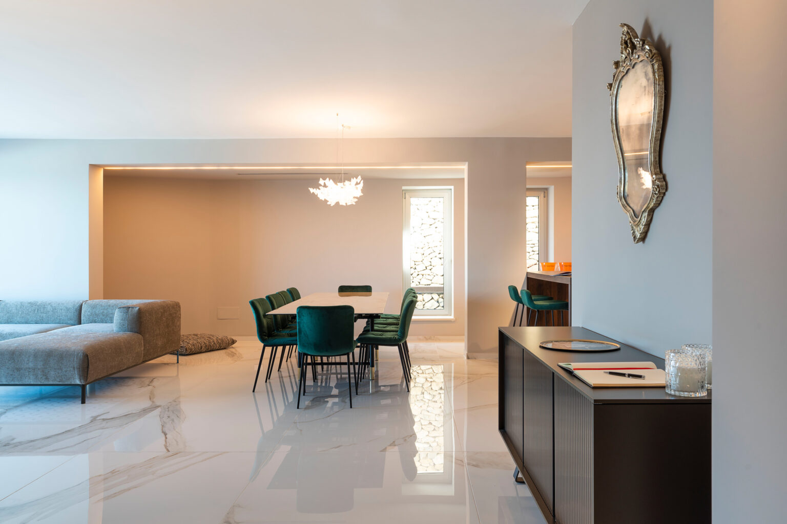 Modern living room and dining area featuring a sleek design, marble flooring, a comfortable sofa, a stylish dining table with green chairs, and an elegant chandelier, complemented by a decorative mirror on the wall. | Only on SopranoVillas