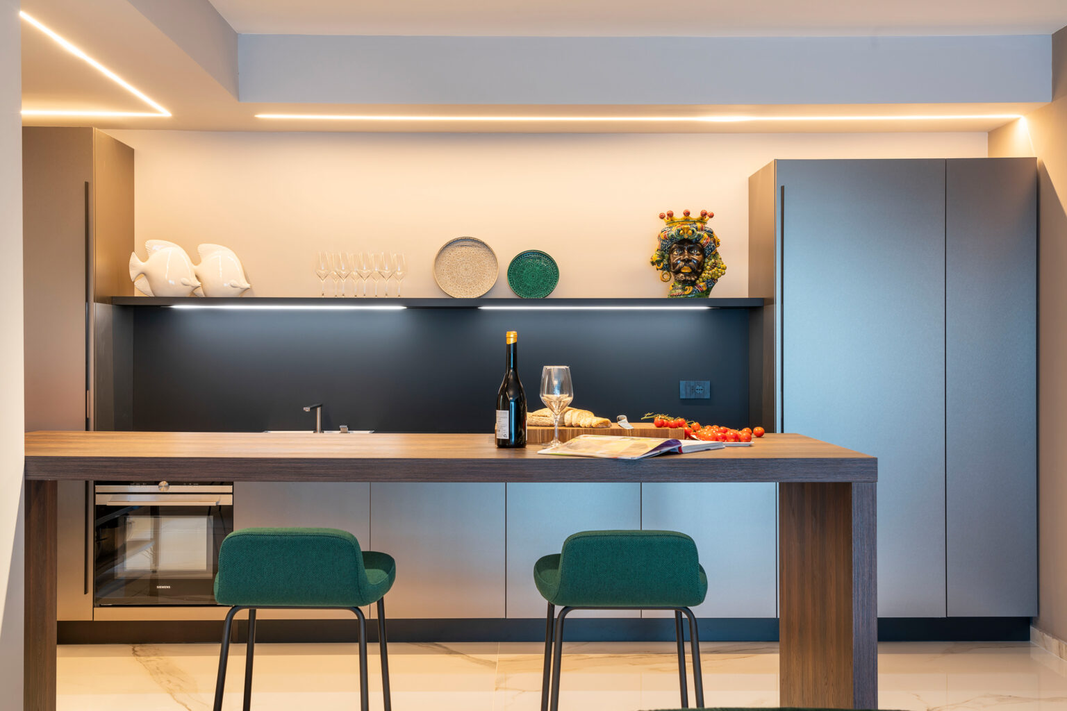 Modern kitchen interior featuring sleek cabinetry, a stylish countertop, and two green chairs. The space is illuminated with warm lighting, showcasing a decorative arrangement of plates and a floral centerpiece, complemented by a bottle of wine and glasses on the counter. | Only on SopranoVillas
