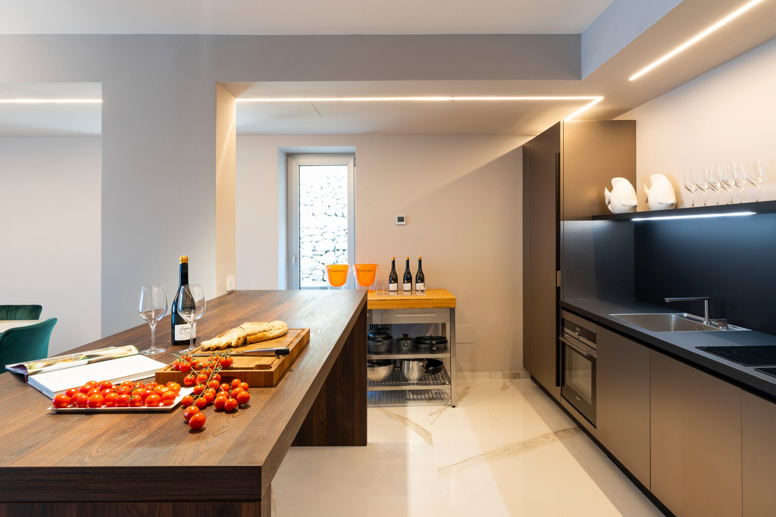 Modern kitchen interior featuring a wooden countertop with fresh tomatoes and a cheese platter, a sleek black cabinetry setup, wine glasses, and a wine rack, illuminated by stylish LED lighting. | Only on SopranoVillas