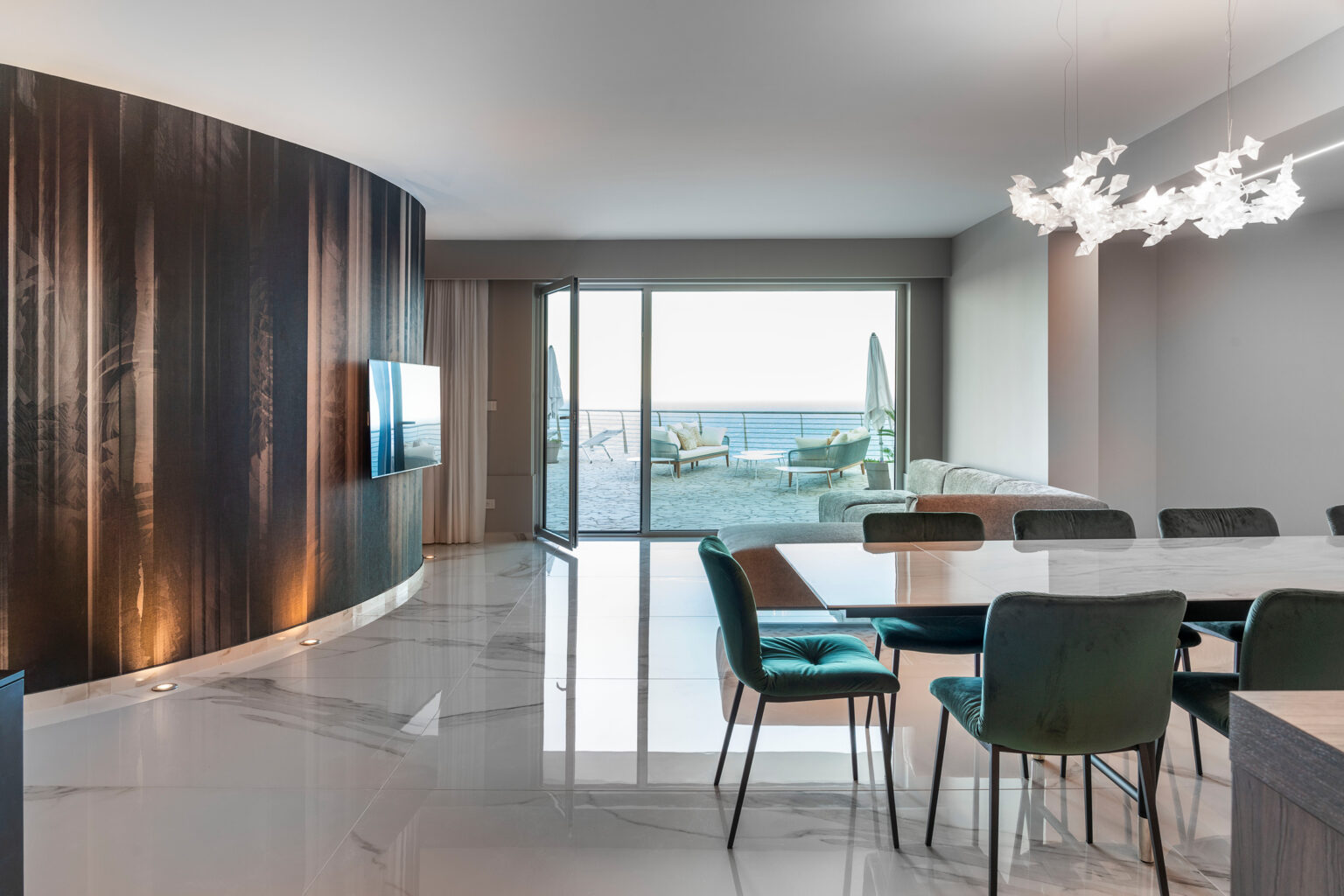 Modern dining area featuring a sleek design with a marble floor, a large dining table surrounded by stylish chairs, and a wall-mounted TV. Large windows offer a view of the outdoor landscape, complemented by contemporary lighting fixtures. | Only on SopranoVillas