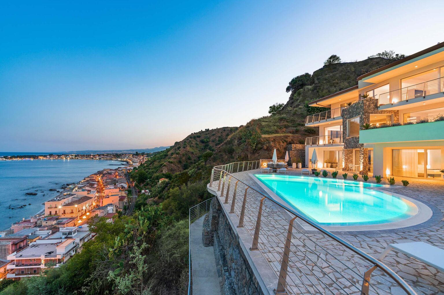 Luxury villa with a stunning infinity pool overlooking the coastline at sunset, featuring modern architecture and scenic views of the ocean and nearby town. | Only on SopranoVillas
