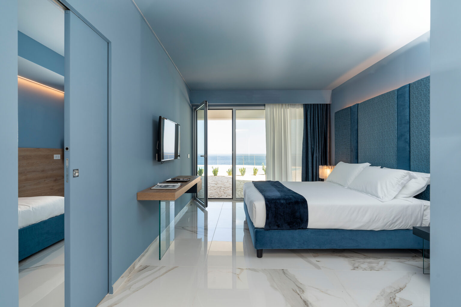 Modern hotel room with blue walls, a king-sized bed, and a flat-screen TV. Features large sliding glass doors leading to a balcony with ocean views. Stylish furnishings include a wooden desk and elegant lighting, complemented by polished marble flooring. Ideal for a relaxing getaway. | Only on SopranoVillas