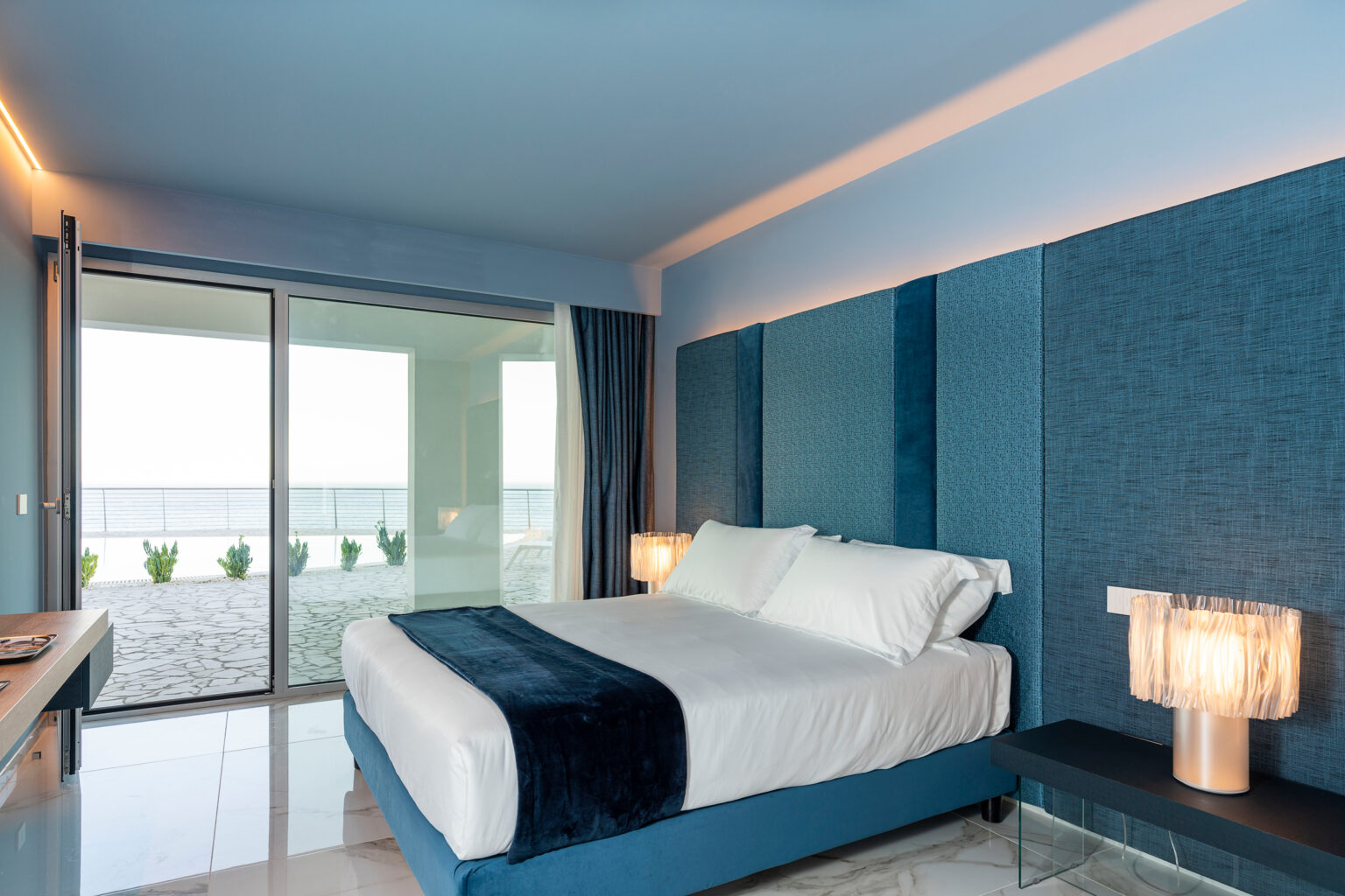 Modern beachfront hotel room featuring a king-sized bed with blue accents, large windows offering ocean views, and stylish lighting. Ideal for a relaxing getaway. | Only on SopranoVillas