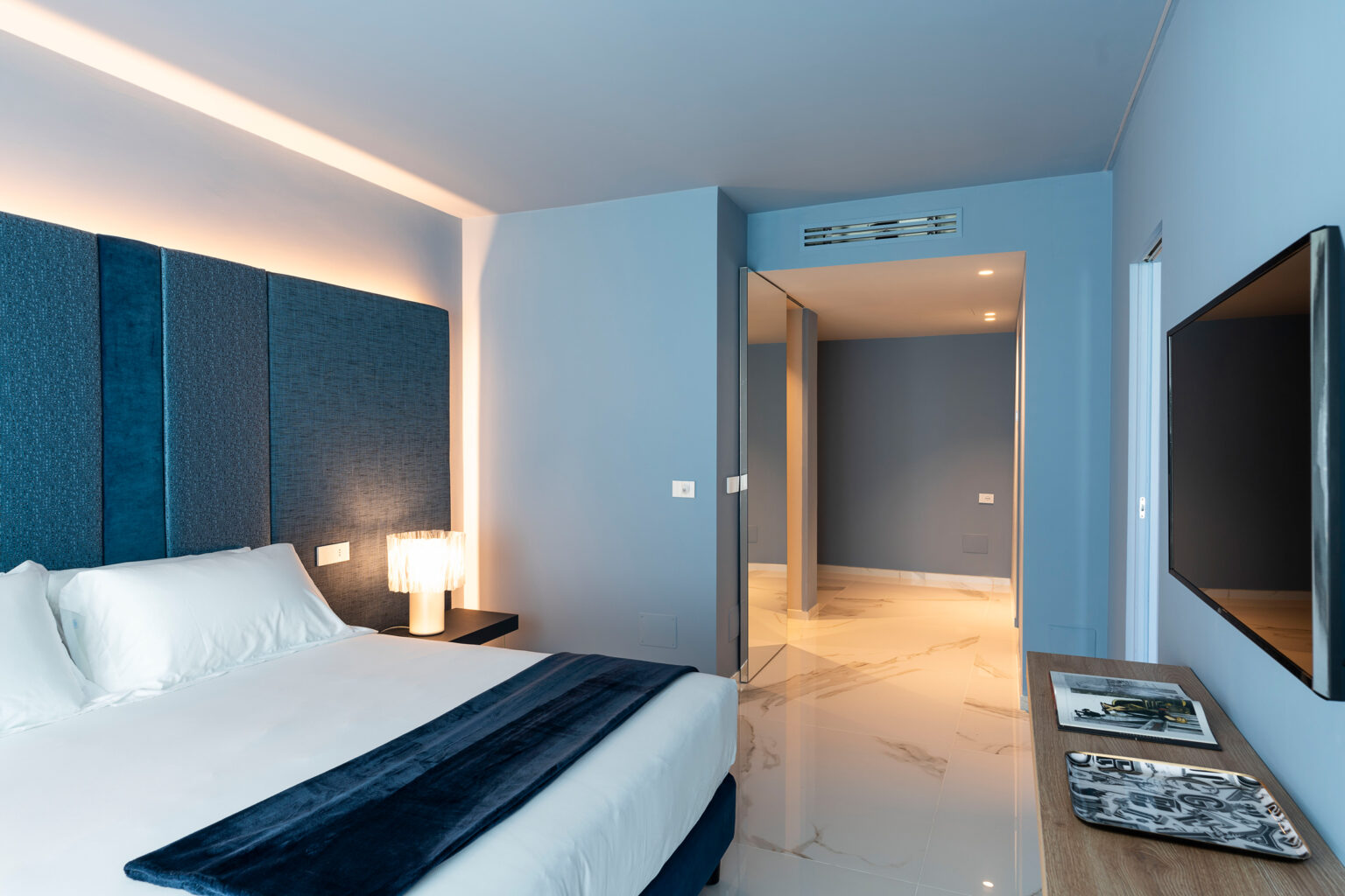 Modern bedroom featuring a blue accent wall, stylish bedding, and a flat-screen TV, with an adjacent doorway leading to a well-lit hallway. The room showcases contemporary design elements and elegant lighting. | Only on SopranoVillas