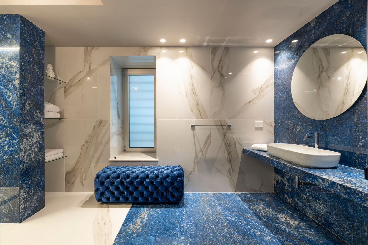 Modern bathroom design featuring blue marble walls, a sleek white countertop with a modern sink, a round mirror, and a stylish blue upholstered bench. Natural light enters through a window, enhancing the luxurious ambiance. | Only on SopranoVillas