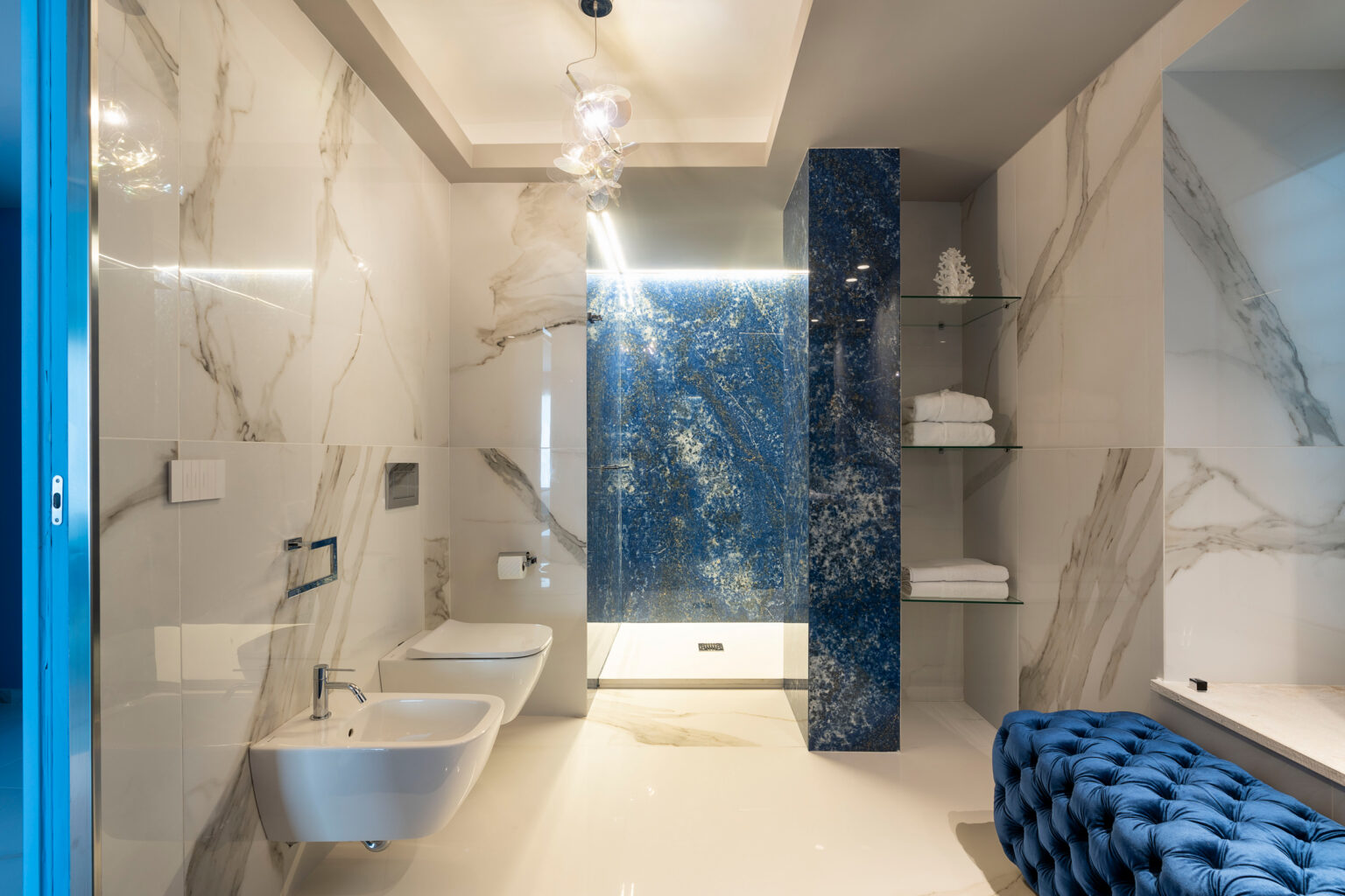 Modern bathroom design featuring elegant marble walls, a blue accent wall with artistic tiles, a contemporary sink and bidet, and stylish lighting. The space is complemented by plush towels and a chic blue upholstered bench. | Only on SopranoVillas