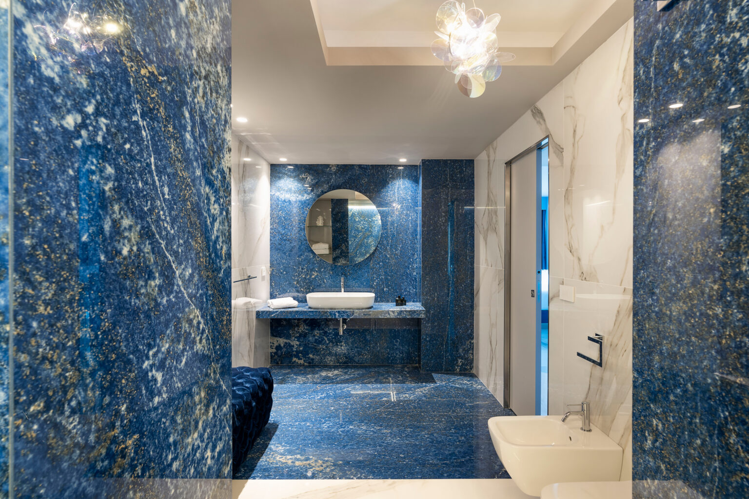 Luxurious modern bathroom featuring striking blue marble walls and flooring, a stylish round mirror above a sleek vanity, and elegant lighting fixtures. | Only on SopranoVillas