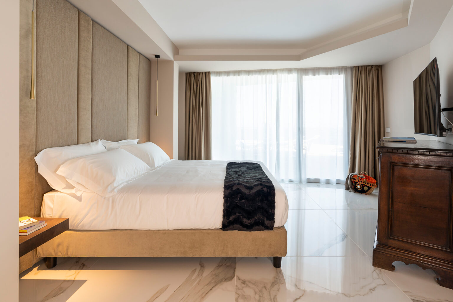 Modern hotel bedroom featuring a king-sized bed with elegant bedding, large windows allowing natural light, and stylish decor, including a wooden dresser and decorative plants. | Only on SopranoVillas