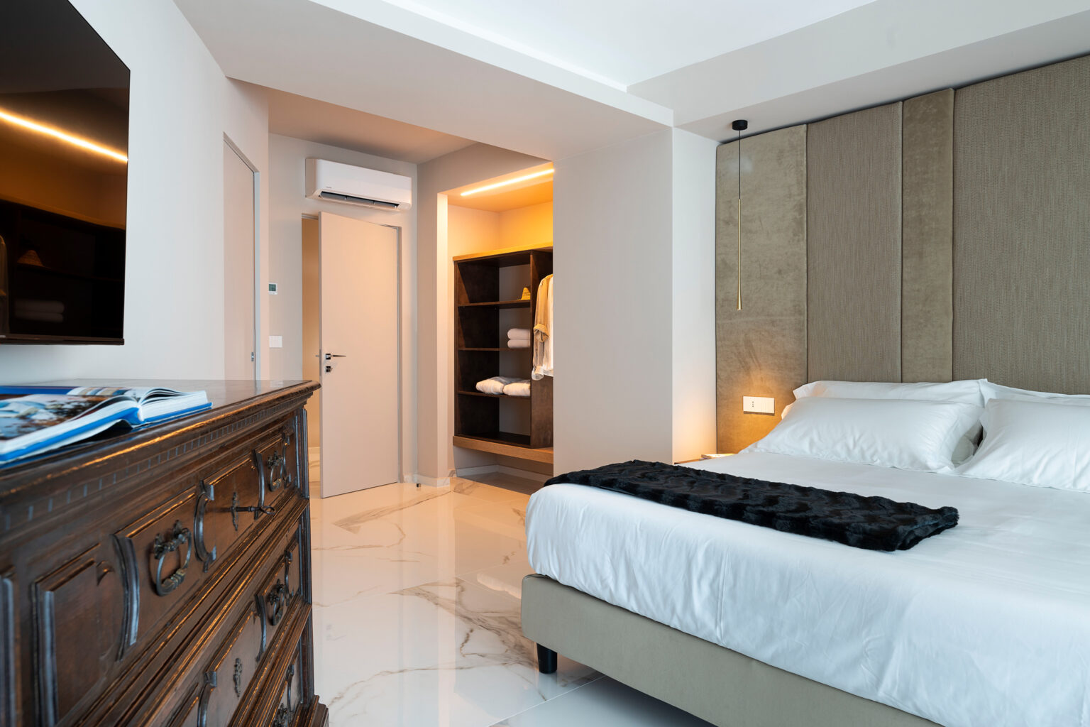 Modern hotel room featuring a comfortable bed with stylish bedding, a wooden dresser, and a well-lit entrance to a bathroom area. The decor combines contemporary elements with elegant finishes, creating a cozy atmosphere. | Only on SopranoVillas