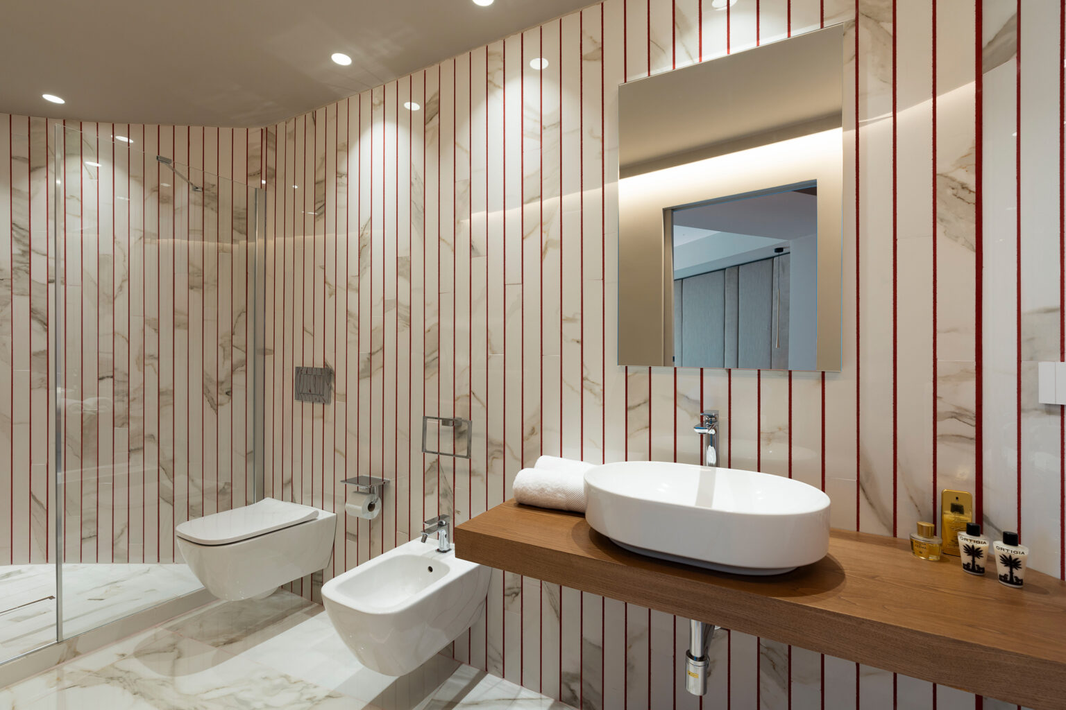 Modern bathroom design featuring a stylish oval sink on a wooden countertop, a bidet, and a large mirror. The walls are adorned with elegant marble and vertical red stripes, complemented by soft lighting for a warm ambiance. | Only on SopranoVillas