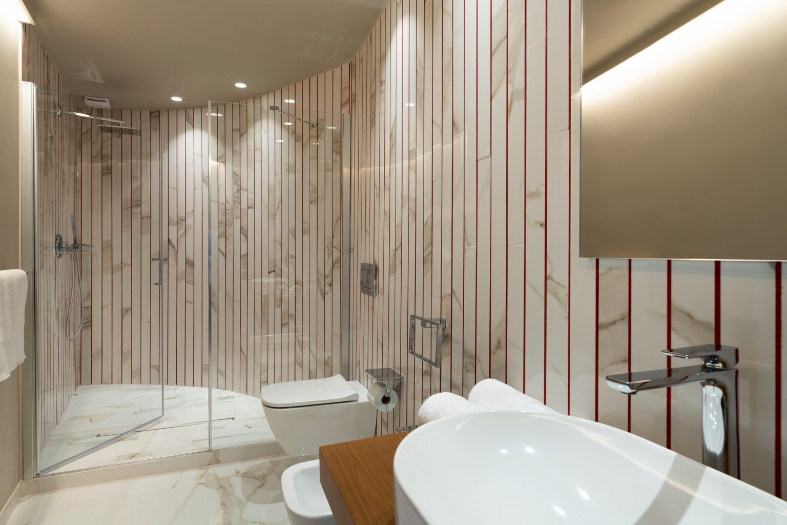 Modern bathroom featuring a spacious glass shower, elegant marble walls with red accents, a sleek toilet, and a stylish sink. Bright lighting enhances the contemporary design. | Only on SopranoVillas