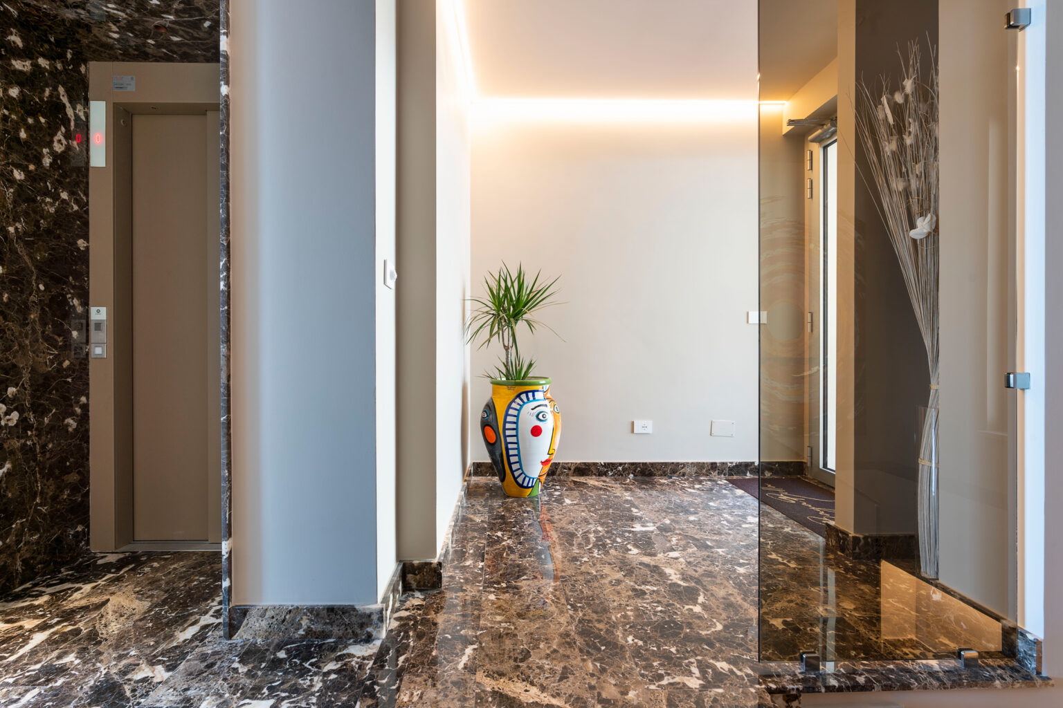 Modern hallway featuring an elevator, marble flooring, and a decorative plant in a colorful vase. The design includes sleek glass doors and contemporary lighting, creating a stylish entrance. | Only on SopranoVillas