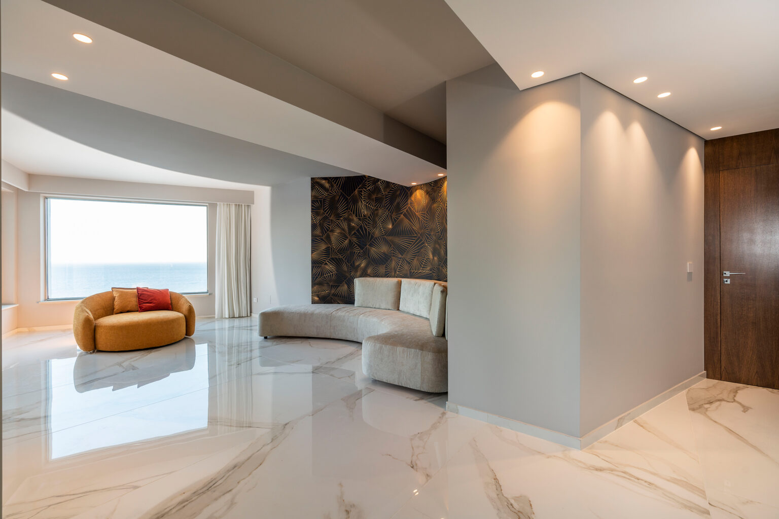 Modern living room interior featuring a curved beige sofa and a circular orange accent chair with a red cushion. The space is illuminated by recessed lighting, showcasing glossy marble flooring and a decorative wall with a tropical leaf pattern. Large window offers a view of the ocean, creating a bright and airy atmosphere. | Only on SopranoVillas