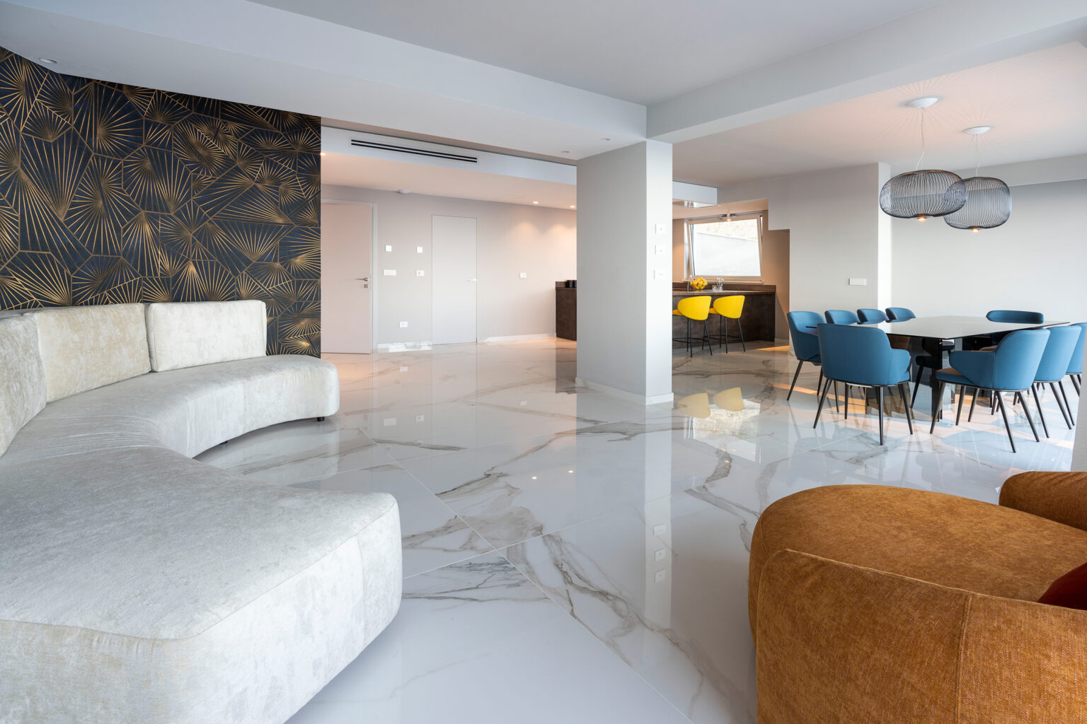 Modern open-concept living space featuring a stylish curved sofa, vibrant blue dining chairs, and a striking gold-patterned accent wall. The polished marble floor reflects natural light, enhancing the contemporary design. | Only on SopranoVillas