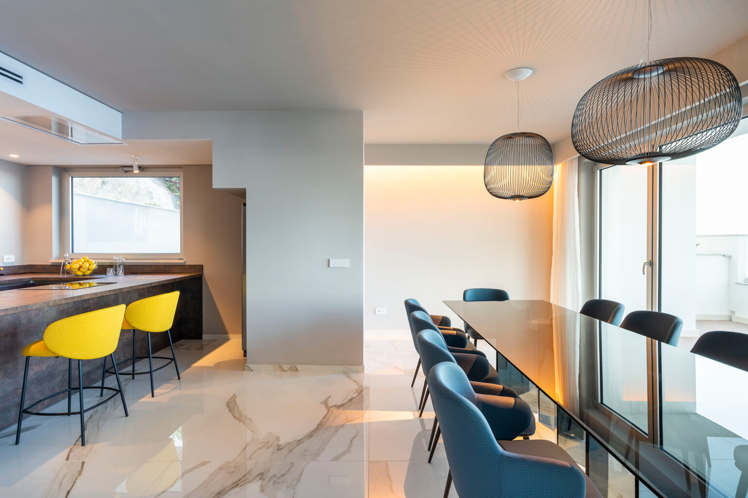 Modern kitchen and dining area featuring a sleek marble floor, a stylish dining table with blue chairs, and yellow bar stools at the kitchen counter. The space is illuminated by contemporary pendant lights and has large windows allowing natural light to fill the room. | Only on SopranoVillas