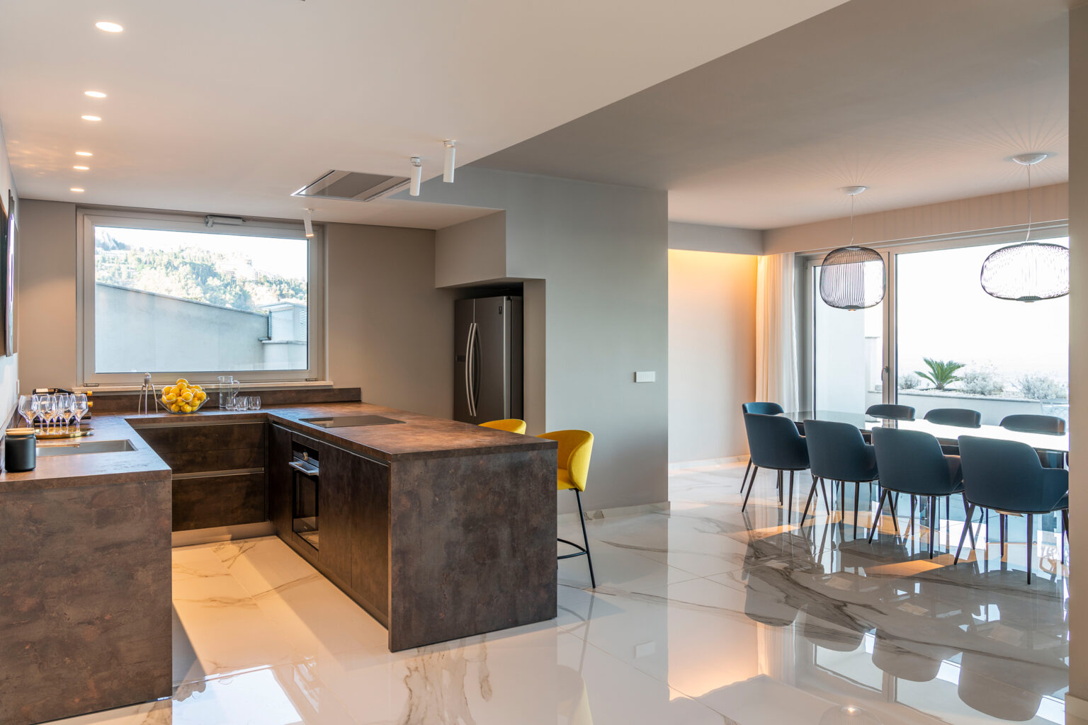 Modern kitchen interior featuring a sleek island with dark countertops, stainless steel appliances, and a large window offering a scenic view. Adjacent dining area with a glass table and blue chairs, illuminated by stylish pendant lights. Elegant marble flooring enhances the contemporary design. | Only on SopranoVillas
