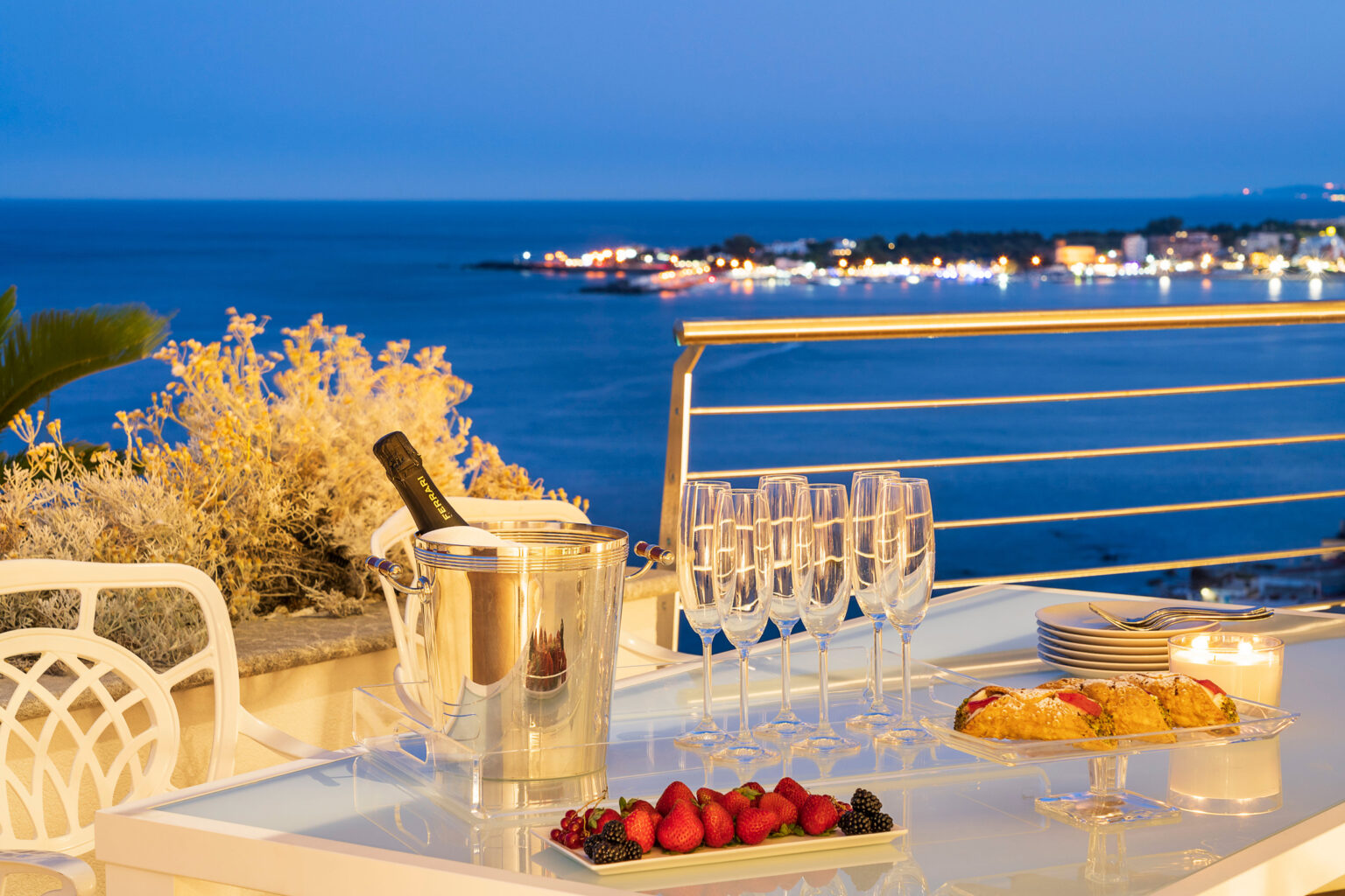 Elegant outdoor dining setup with a stunning ocean view at sunset, featuring a champagne bucket, sparkling glasses, a platter of fresh strawberries and blackberries, and delicious pastries on a glass table, perfect for a romantic evening or celebration. | Only on SopranoVillas