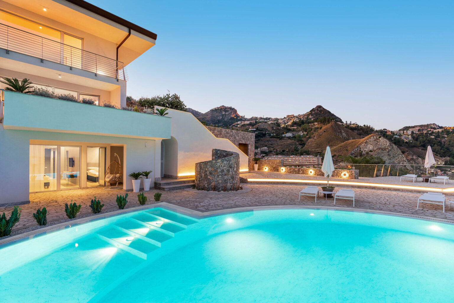 Luxury villa with a modern design, featuring a sparkling pool surrounded by lounge chairs and illuminated landscaping, set against a backdrop of mountains at dusk. | Only on SopranoVillas