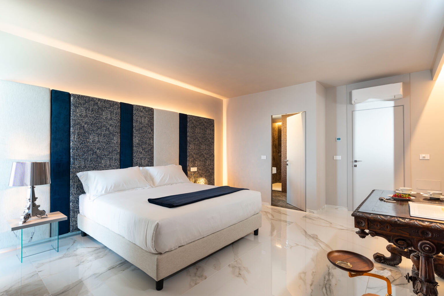 Modern hotel bedroom featuring a plush king-sized bed with white linens, stylish blue accent walls, and elegant marble flooring. The room includes a bedside lamp, a wooden table with refreshments, and a doorway leading to a bathroom, creating a luxurious and inviting atmosphere. | Only on SopranoVillas