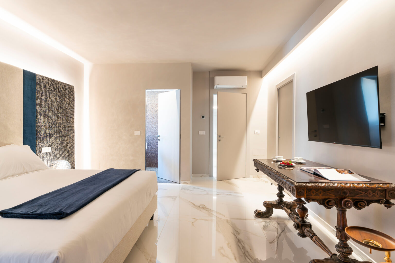 Modern hotel room featuring a plush bed with a navy throw, elegant marble flooring, and a stylish wooden table with refreshments. The room includes a wall-mounted TV, contemporary lighting, and two doors leading to adjoining spaces, showcasing a blend of comfort and sophistication. | Only on SopranoVillas