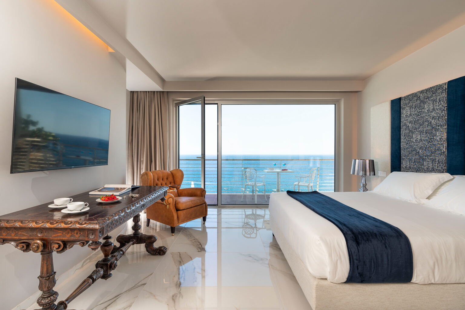 Modern hotel room with ocean view, featuring a king-sized bed, stylish armchair, wooden table with refreshments, and large glass doors opening to a balcony. Elegant decor and natural light create a relaxing atmosphere. | Only on SopranoVillas