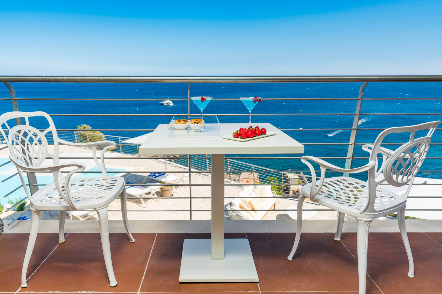 Cozy balcony dining setup with a table and two chairs overlooking the ocean, featuring a plate of strawberries and cocktails, ideal for a relaxing seaside experience. | Only on SopranoVillas