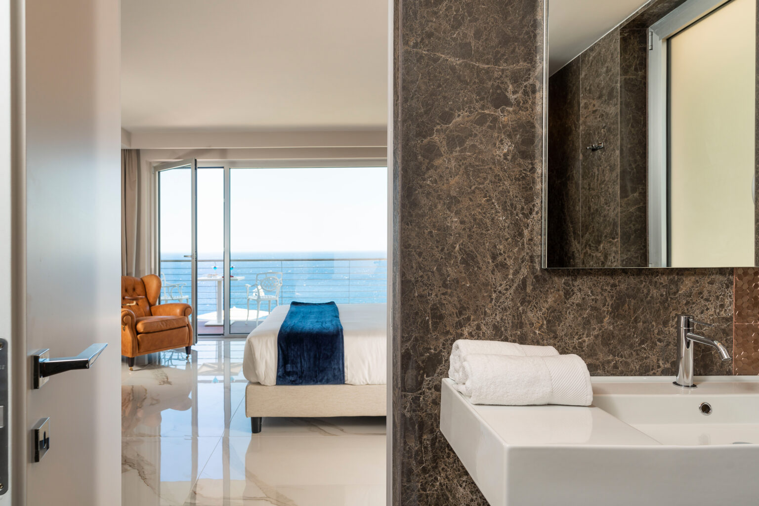 Modern hotel room with a stunning ocean view, featuring a stylish bathroom with a sink and mirror, and a cozy seating area with an armchair. | Only on SopranoVillas