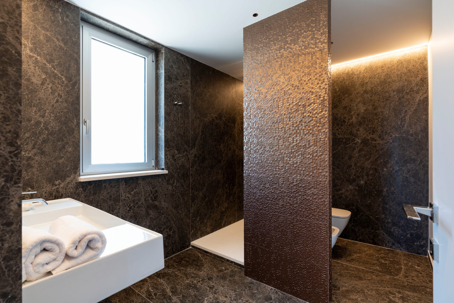 Modern bathroom interior featuring dark stone walls, a sleek white sink with rolled towels, and a stylish partition. Natural light enters through a window, enhancing the contemporary design. | Only on SopranoVillas