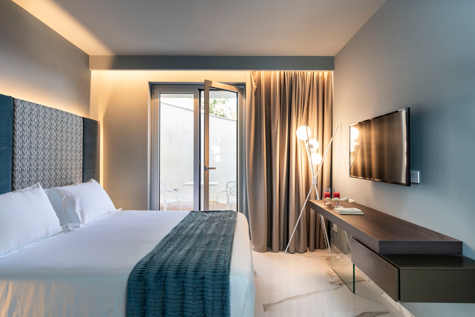 Modern hotel room featuring a king-sized bed with white linens, a stylish gray accent wall, a flat-screen TV, a sleek wooden desk, and large windows with sheer curtains. The design includes contemporary lighting and a minimalist aesthetic, creating a comfortable and inviting atmosphere. | Only on SopranoVillas